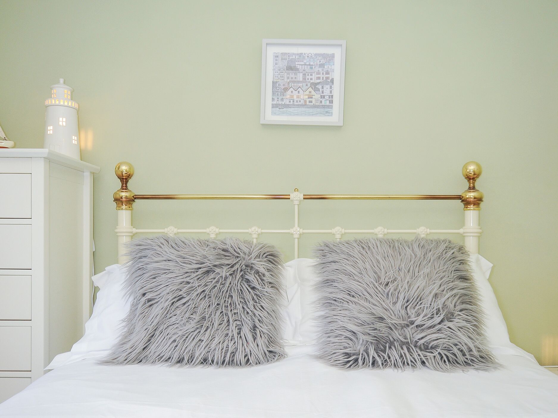 Un lit avec une tête de lit blanche et deux oreillers moelleux, une commode blanche et un tableau encadré au mur.