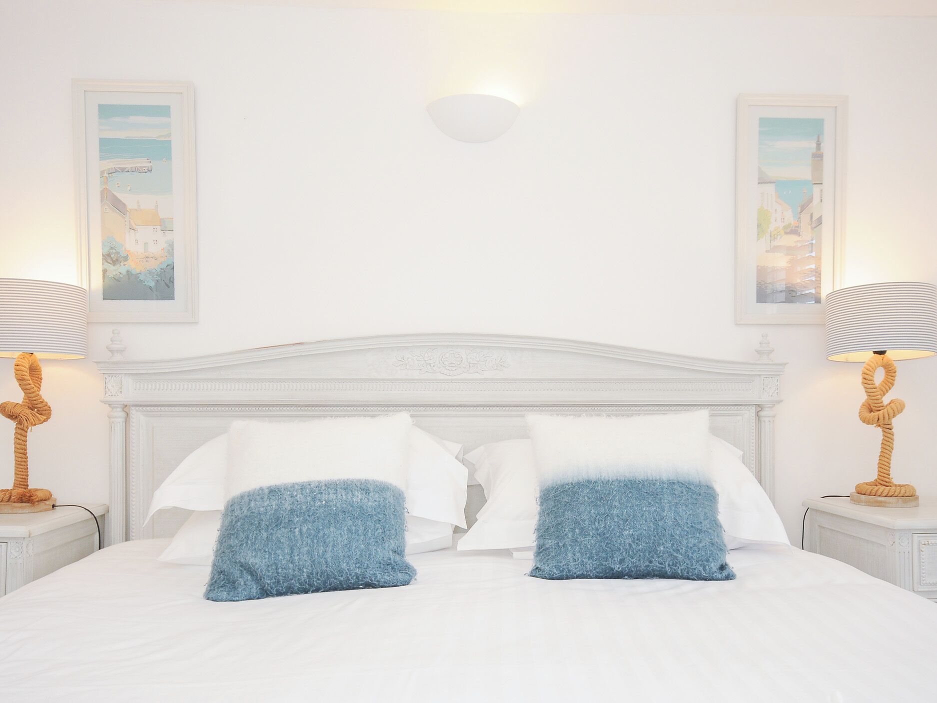 Een net opgemaakt bed met witte en blauwe kussens, een wit hoofdbord en twee wandlampen.