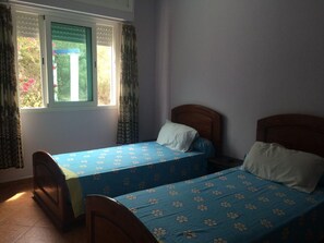 A room with two single beds, a window with curtains, and a view of greenery outside.