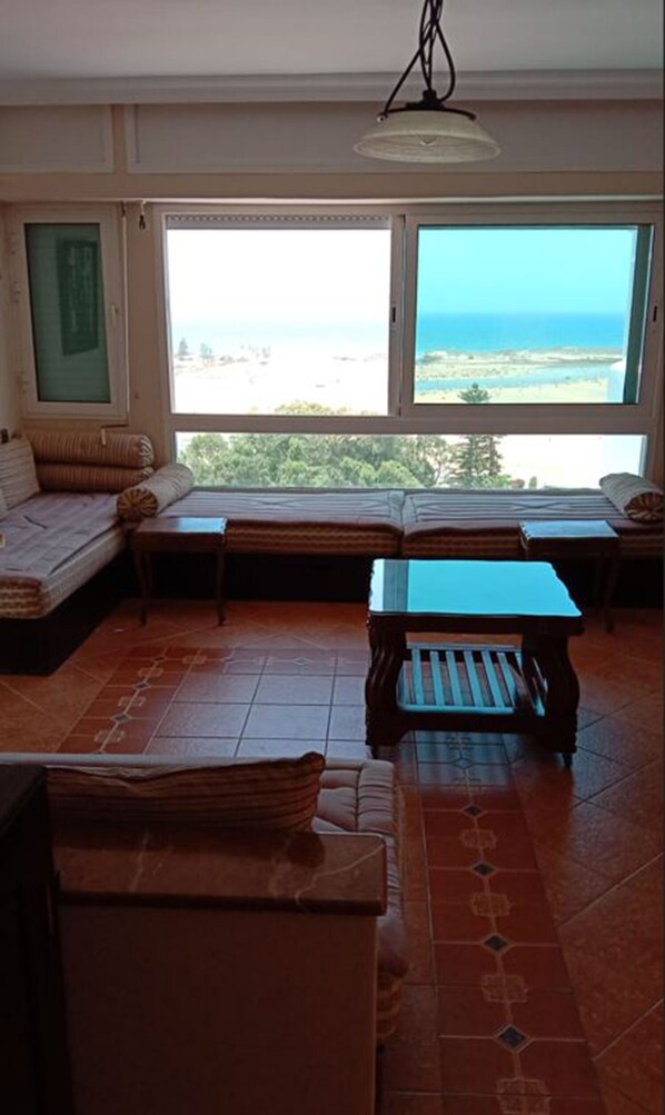 A room with a large window offering a view of the beach and ocean. There is a sofa and a small table in the room.