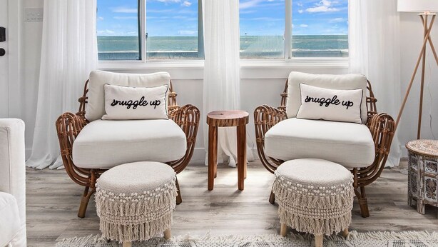 A room with two wicker chairs, each with a cushion and a pillow that says "snuggle up". There is a small round wooden table between the chairs. The room has white curtains and a view of the ocean through large windows. - Lux Beachfront House*Private*Fully Loaded*Firepit (Gulf Shores)