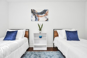 A hotel room with two beds, a white wall, a blue abstract painting, a small white shelf with a plant, and a blue rug. - ✵Hidden Gem Safari✵| Home w/ Backyard ⮕ Midtown (Houston)