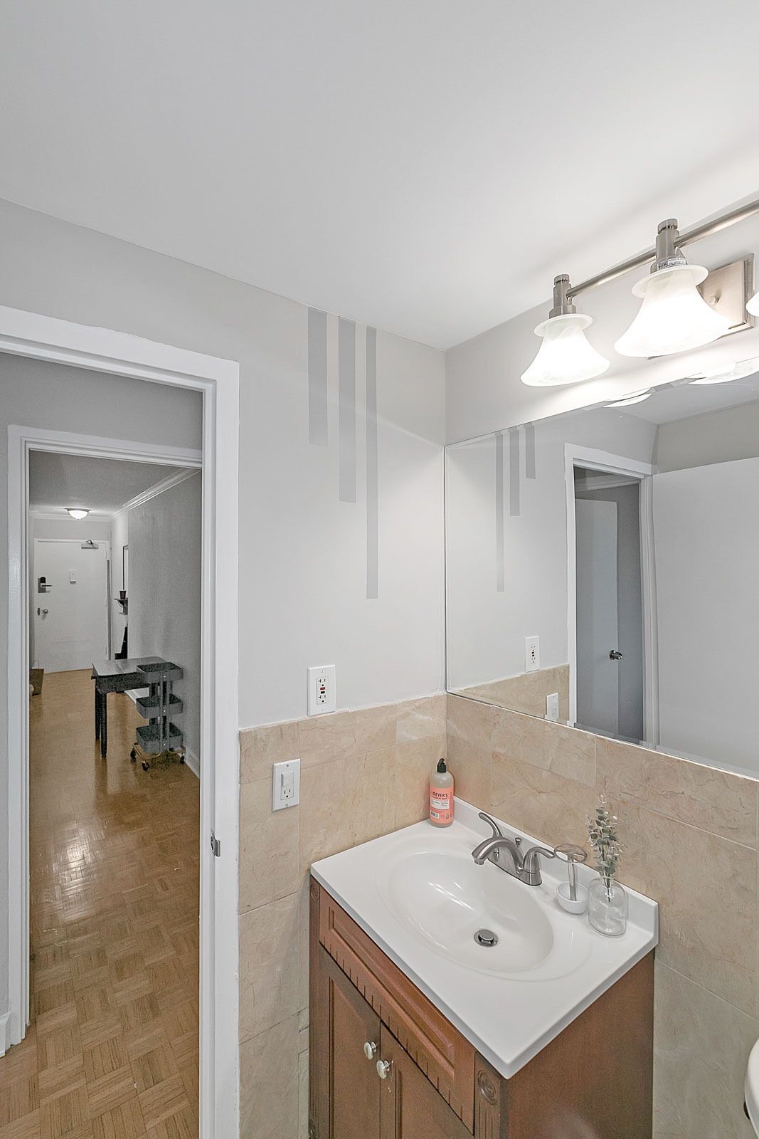 A bathroom with a white sink, a mirror, and a light fixture above it. There is a bottle of hand soap on the counter.