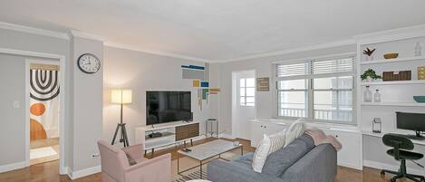 Un salon moderne avec un canapé gris, un fauteuil rose, une table basse et un téléviseur posé sur un meuble blanc.