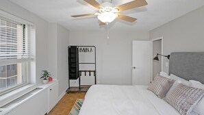 A bedroom with a bed, a ceiling fan, a window with blinds, a radiator, a shelf with plants, and a door leading to another room. - 21 first floor cozy condo in the center of DT Atlanta (Atlanta)