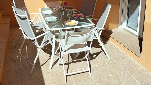 Eine Terrasse mit Tisch für zwei Personen, Stühlen und einem gestreiften Sonnensegel.