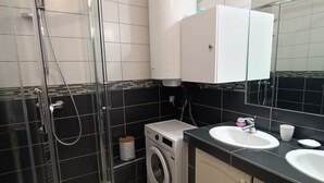Ein Badezimmer mit einer Duschkabine, einer Waschmaschine, einem Waschbecken mit Spiegel und gefliesten Wänden.