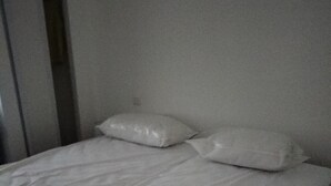 Ein Bett mit weißer Bettwäsche und zwei Kissen in einem Zimmer mit schlichten Wänden und einem Fliesenboden.