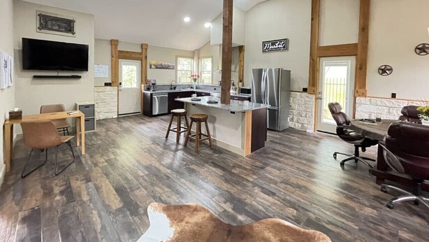Una cocina amplia con una isla central, taburetes de madera, un comedor con mesa y sillas, y un televisor grande montado en la pared.