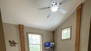 Un dormitorio con una cama, una silla, un televisor y un ventilador de techo.