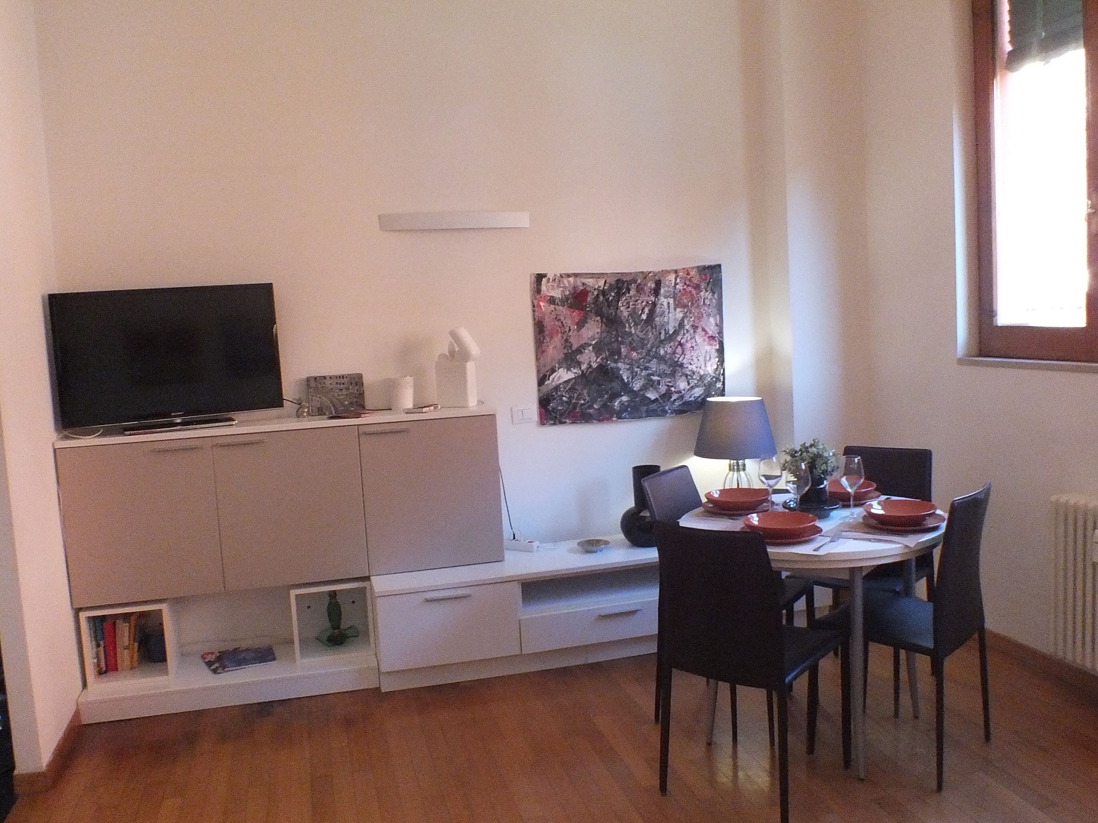 A compact living space with a flat-screen TV, a dining table with chairs, a lamp, and a painting on the wall.