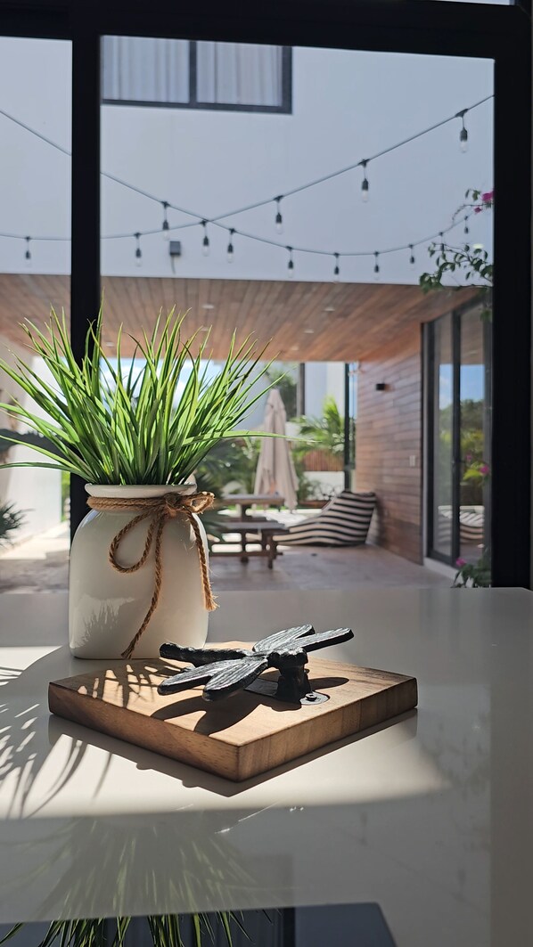 Un interior moderno con una planta en maceta sobre una mesa, un adorno metálico y vistas a un patio exterior con luces colgantes.