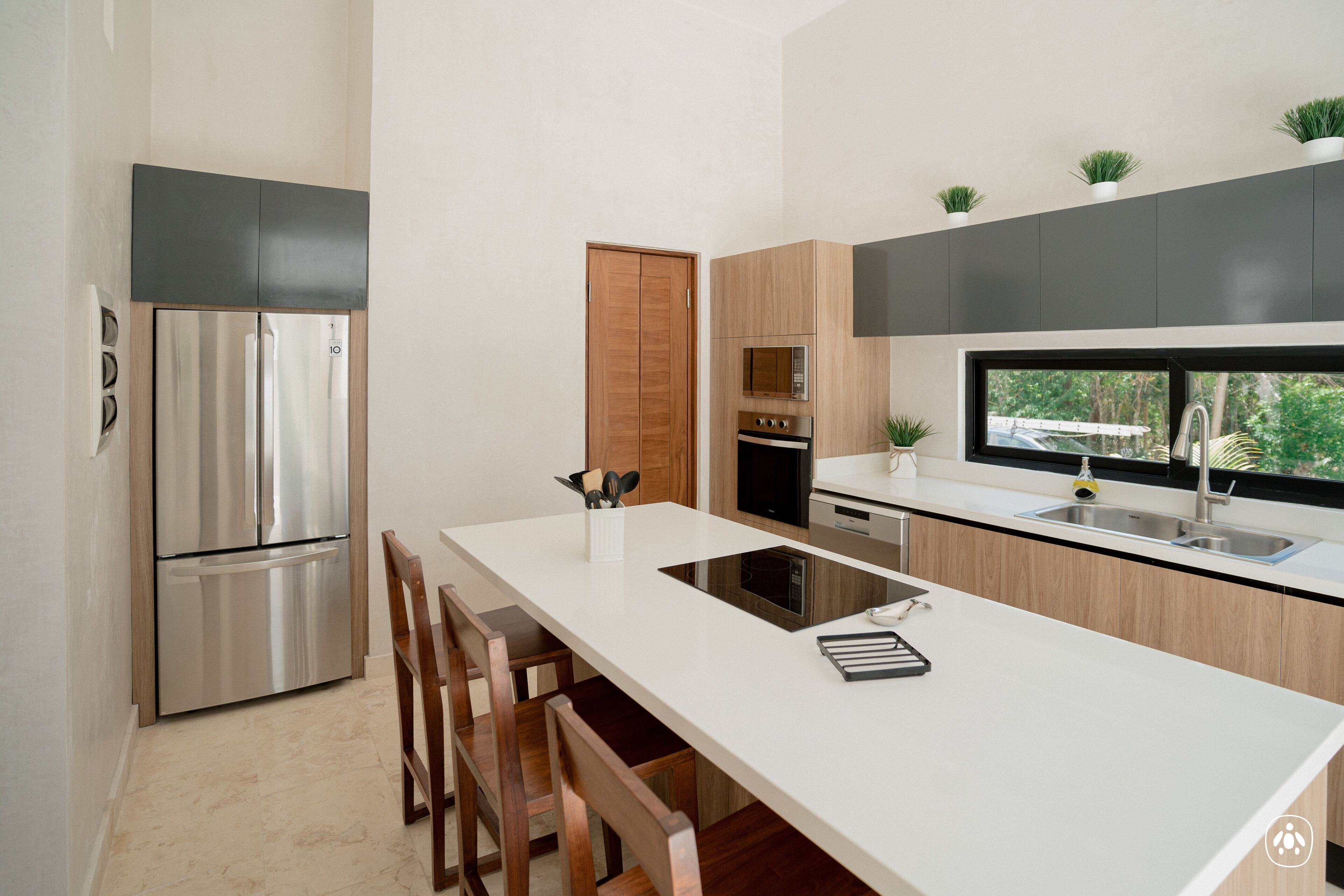 Eine moderne Küche mit einem Edelstahl-Kühlschrank, Holzschränken, einer weißen Arbeitsplatte und einem Fenster mit Blick auf Grünflächen.