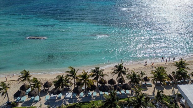 Een strand met helder turquoise water, palmbomen en ligbedden.