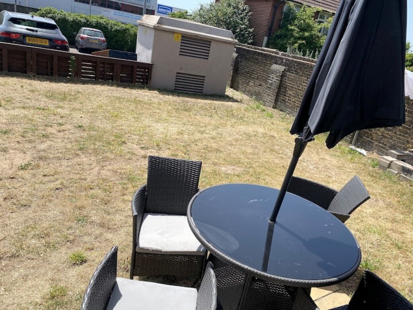 Ein Sitzbereich im Freien mit einem runden Tisch, zwei Stühlen, einem schwarzen Sonnenschirm und einer Backsteinmauer im Hintergrund.