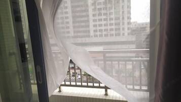 A view from a balcony with a white curtain partially drawn, overlooking a street and buildings.