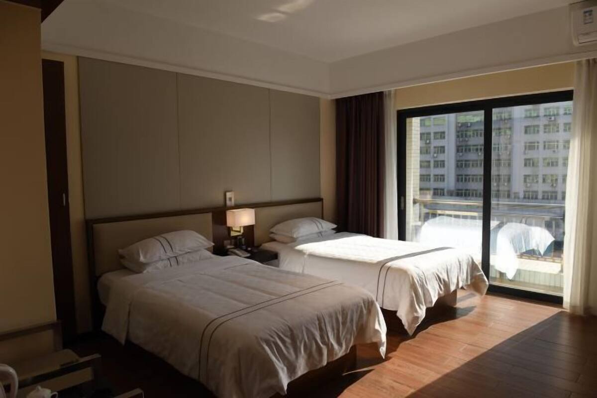 A hotel room with two beds, a nightstand with a lamp, a large window with a view of buildings, and a wooden floor.