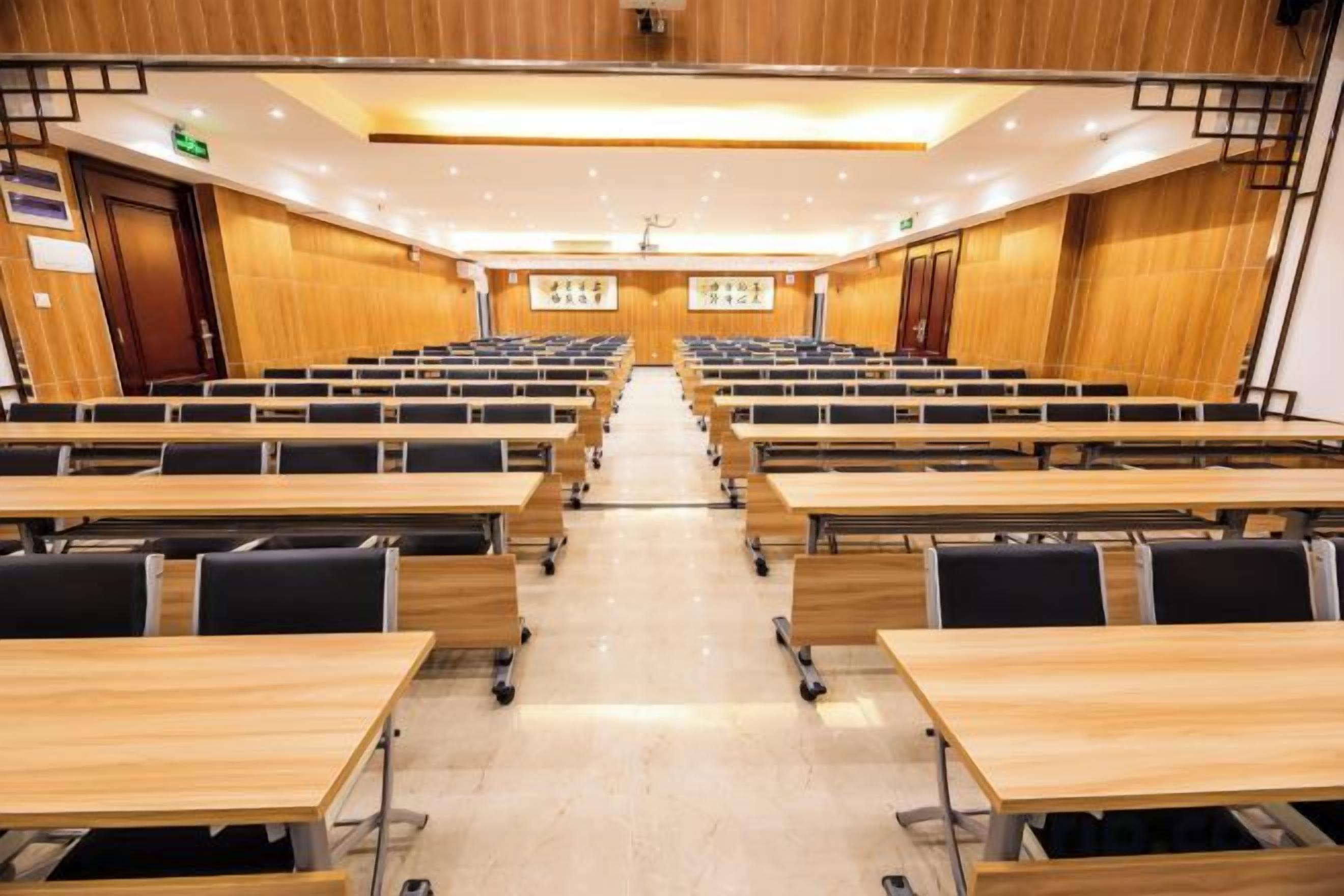 Un salón de conferencias con filas de mesas y sillas, suelo de madera e iluminación empotrada en el techo.