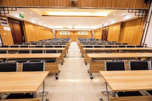 A conference room with rows of tables and chairs, a wooden floor, and a ceiling with recessed lighting.