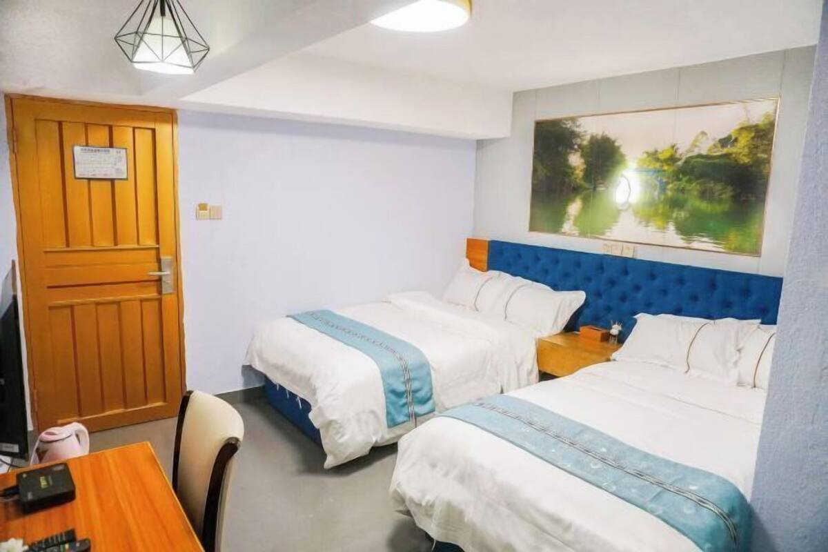 A hotel room with two beds, a wooden door, a desk with a computer, and a large window with a painting.