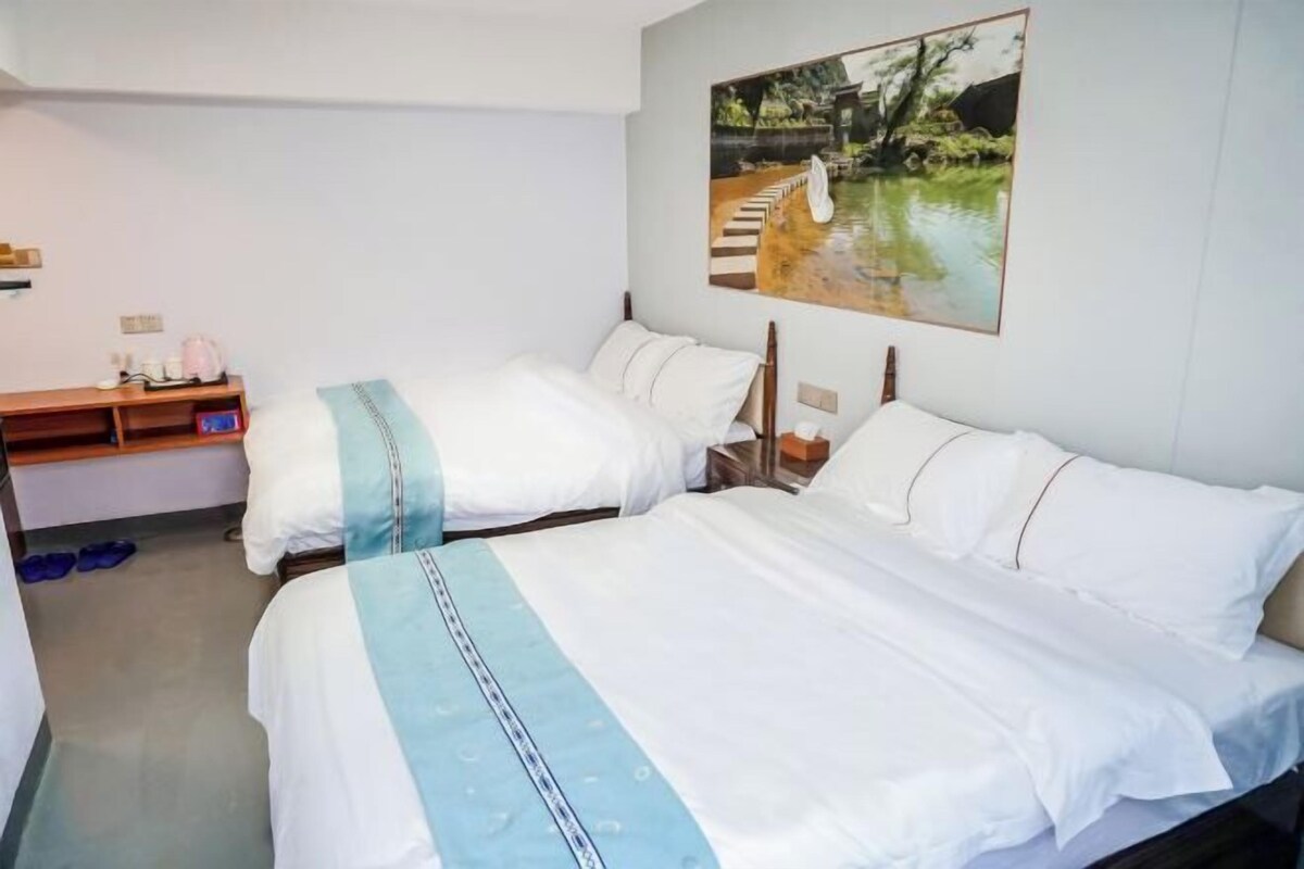 A hotel room with two beds, a wooden nightstand, a small table with a lamp, and a painting of a landscape on the wall.