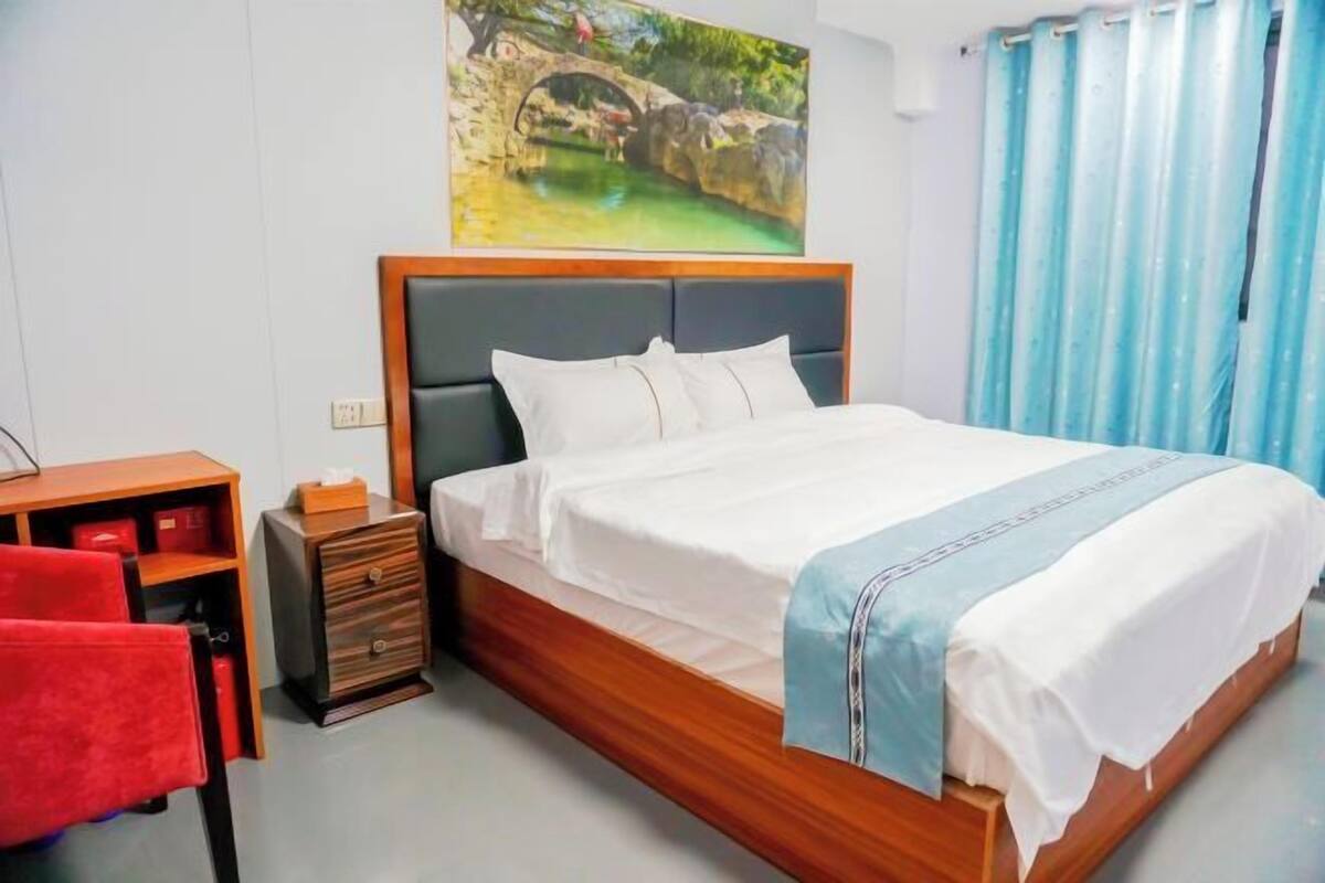 A neatly made bed with a wooden headboard, a small bedside table with drawers, a red chair, and a painting of a bridge over water on the wall.