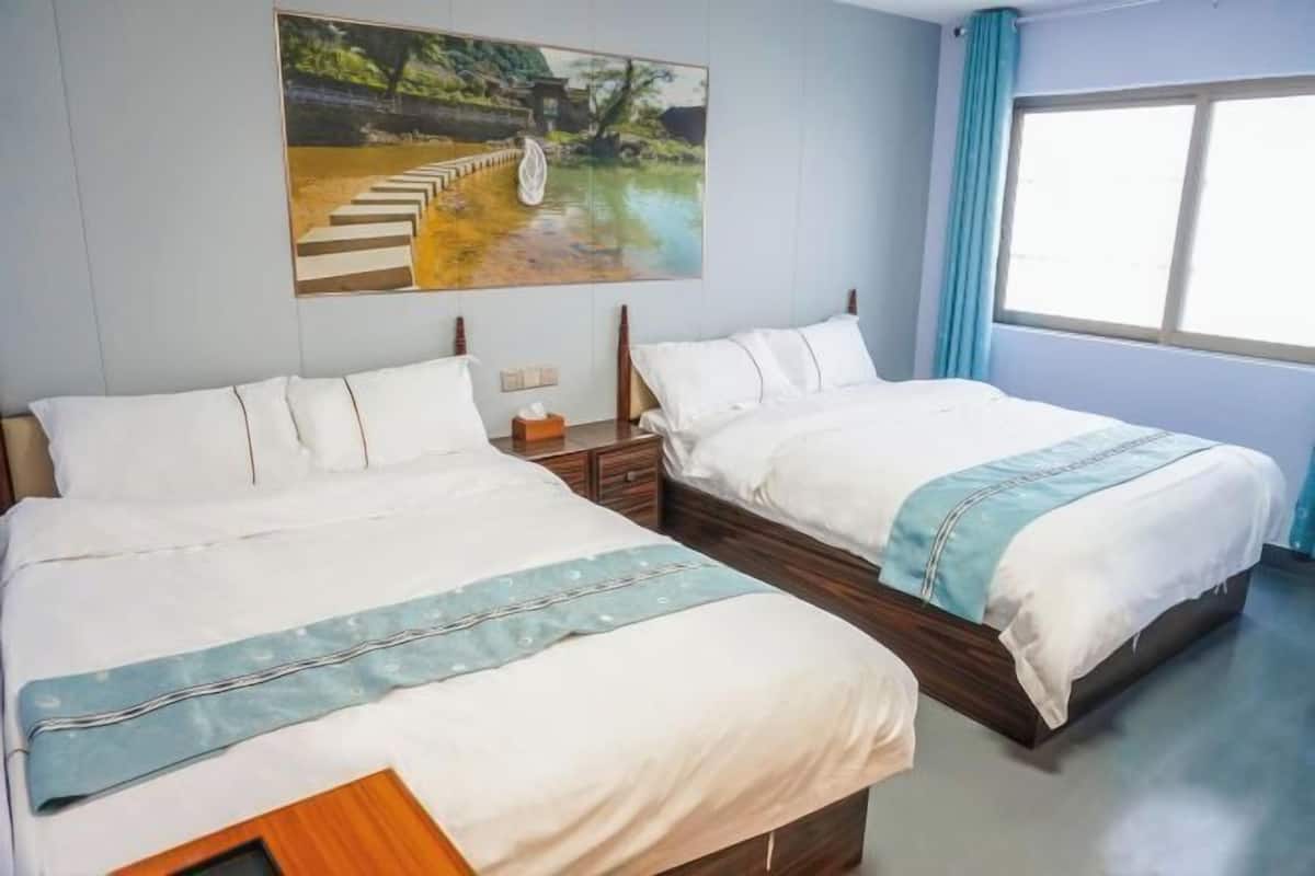 A hotel room with two beds, a painting on the wall, a window with curtains, and a bedside table with a lamp.