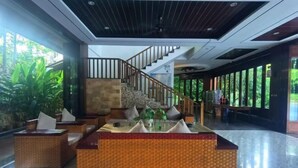 A lobby with a reception desk, seating area, staircase, and large windows with greenery outside.
