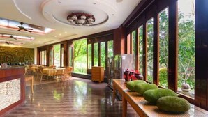 A spacious hotel lobby with a polished wooden floor, large windows offering a view of greenery, and a central seating area with tables and chairs.