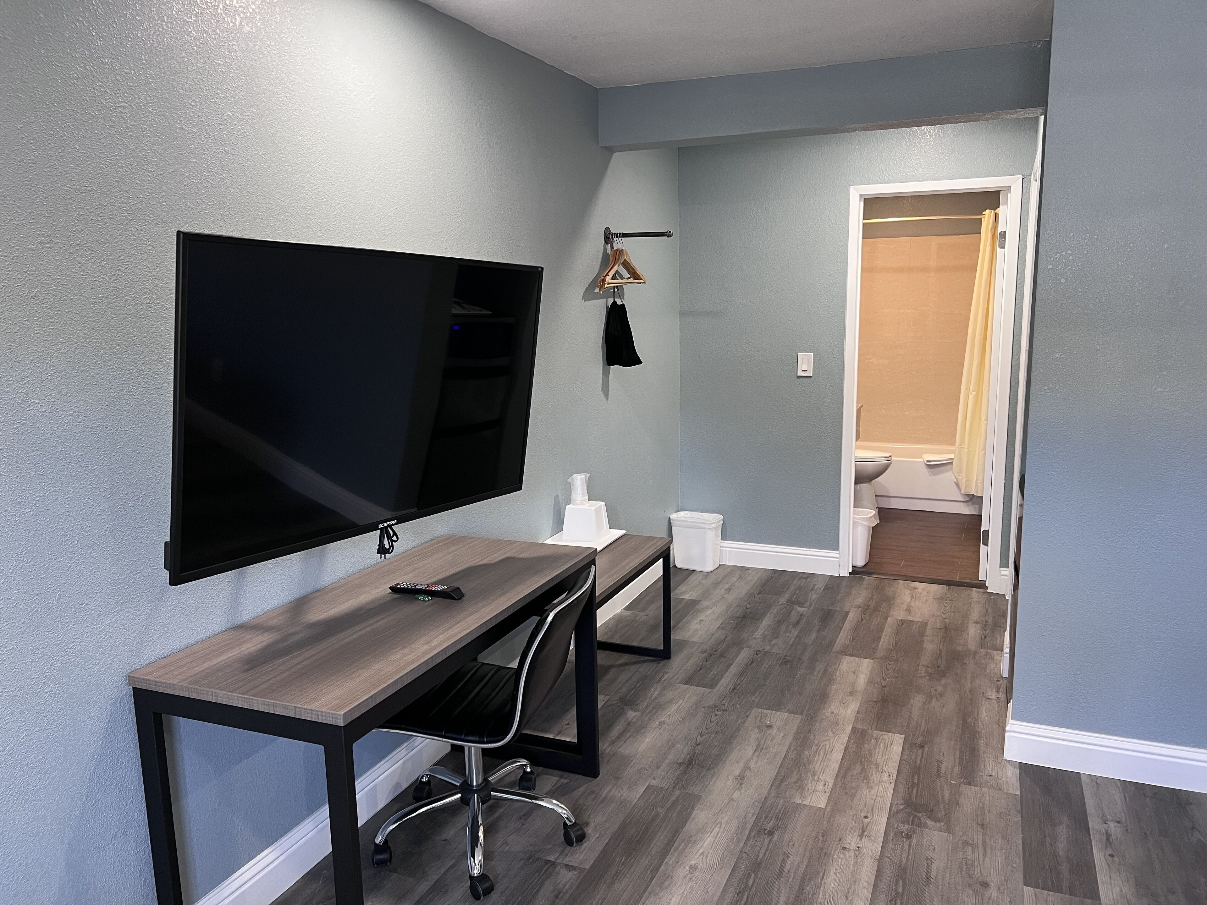 A modern room with a flat-screen TV mounted on the wall, a wooden desk with a chair, and a hallway leading to a bathroom.