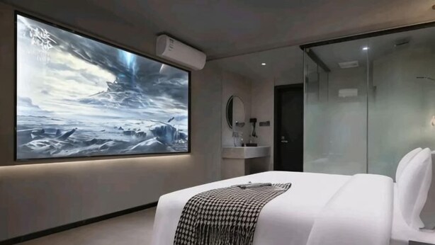 Een moderne hotelkamer met een groot bed, nachtkastje, spiegel en een grote wandtelevisie waarop een landschapsbeeld wordt getoond.
