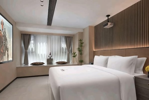 A modern hotel room with a large bed, a window with curtains, a wall-mounted light, and a small table with a vase.