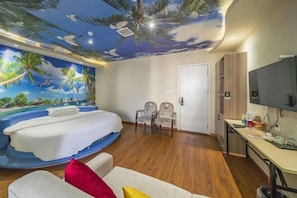 Habitación con un mural de una playa tropical, una cama, dos sillas, un escritorio con televisor y una puerta.
