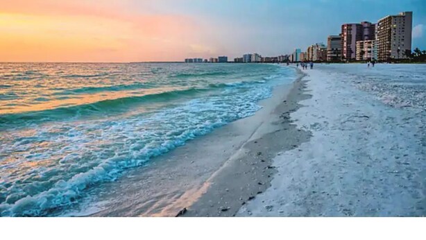 A beach with waves, a clear sky, and buildings in the background.