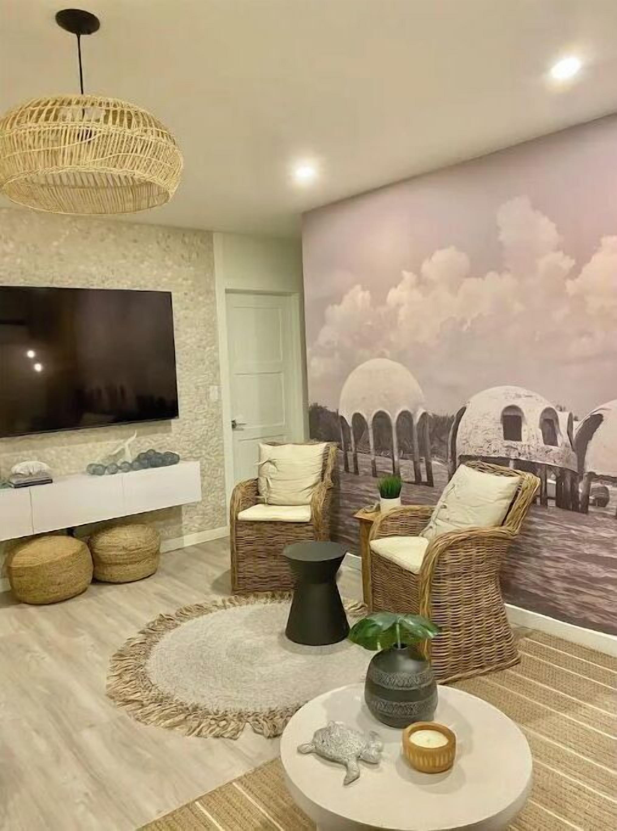 A living room with wicker chairs, a round coffee table, a flat-screen TV, and a mural of a coastal scene.