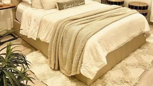 A bedroom with a large bed, a bedside table, a lamp, a wall-mounted decorative item, and a potted plant.