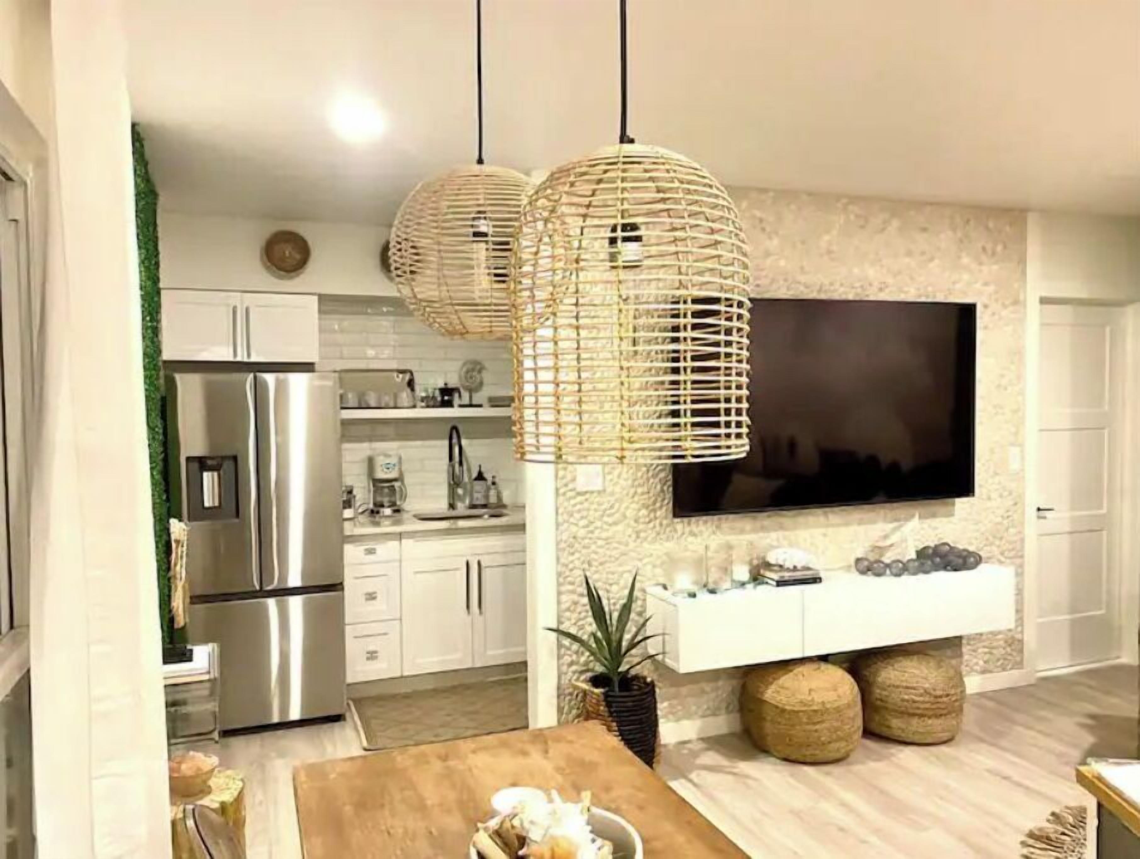 A modern kitchen with stainless steel appliances, a large flat-screen TV mounted on the wall, and two woven pendant lights.