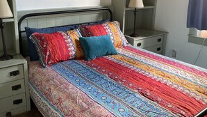 A bedroom with a patterned bedspread, a nightstand with a lamp, a dresser with drawers, a painting on the wall, and a window with a curtain. - Boho Hip Bungalow (Bisbee)
