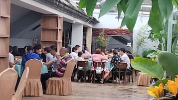 An outdoor dining area with tables and chairs, surrounded by plants and a building with traditional architecture.