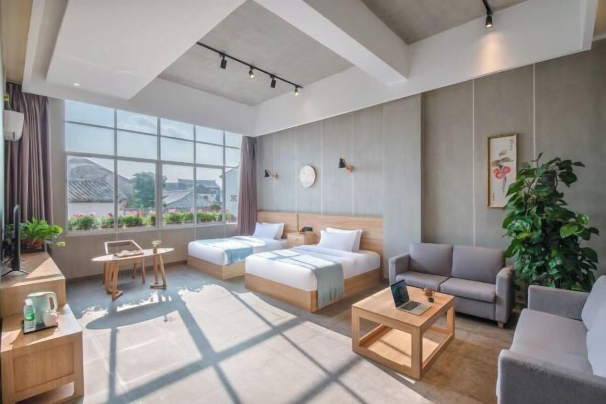 A modern hotel room with two single beds, a sofa, a coffee table, and a large window offering a view of the cityscape.