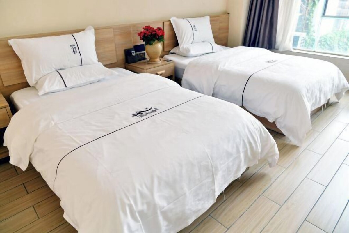 Quarto de hotel com duas camas, uma mesa de cabeceira com um vaso de flores e uma janela com cortinas.