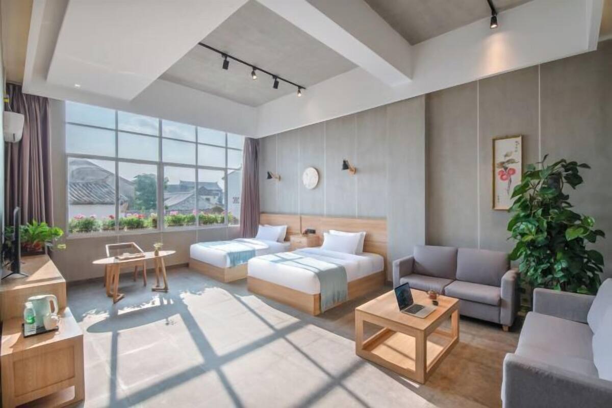 A modern hotel room with two beds, a sofa, a coffee table, and a large window offering a view of the city.