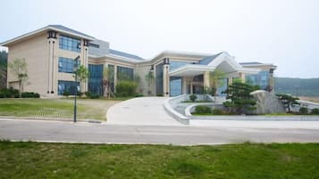 A modern building with a curved driveway, a landscaped lawn, and a body of water in the background.