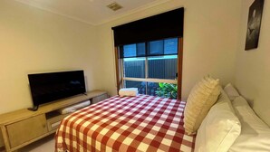 Une chambre avec une couvre-lit à carreaux, un téléviseur posé sur un meuble en bois, une fenêtre avec des stores et une plante près de la fenêtre.