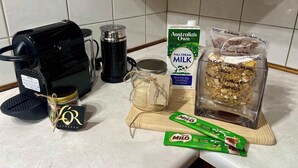Un comptoir de cuisine avec une machine à café, un carton de lait, un pot de sucre, une boîte de céréales et un sachet de noix.