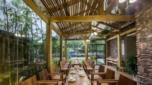A restaurant with a long wooden table set for dining, surrounded by wooden chairs, with a stone wall and glass windows on one side. - Yueming Xiaoyuan Hostel (Wuyishan)