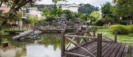 A wooden bridge over a calm water body, surrounded by greenery and buildings in the background.