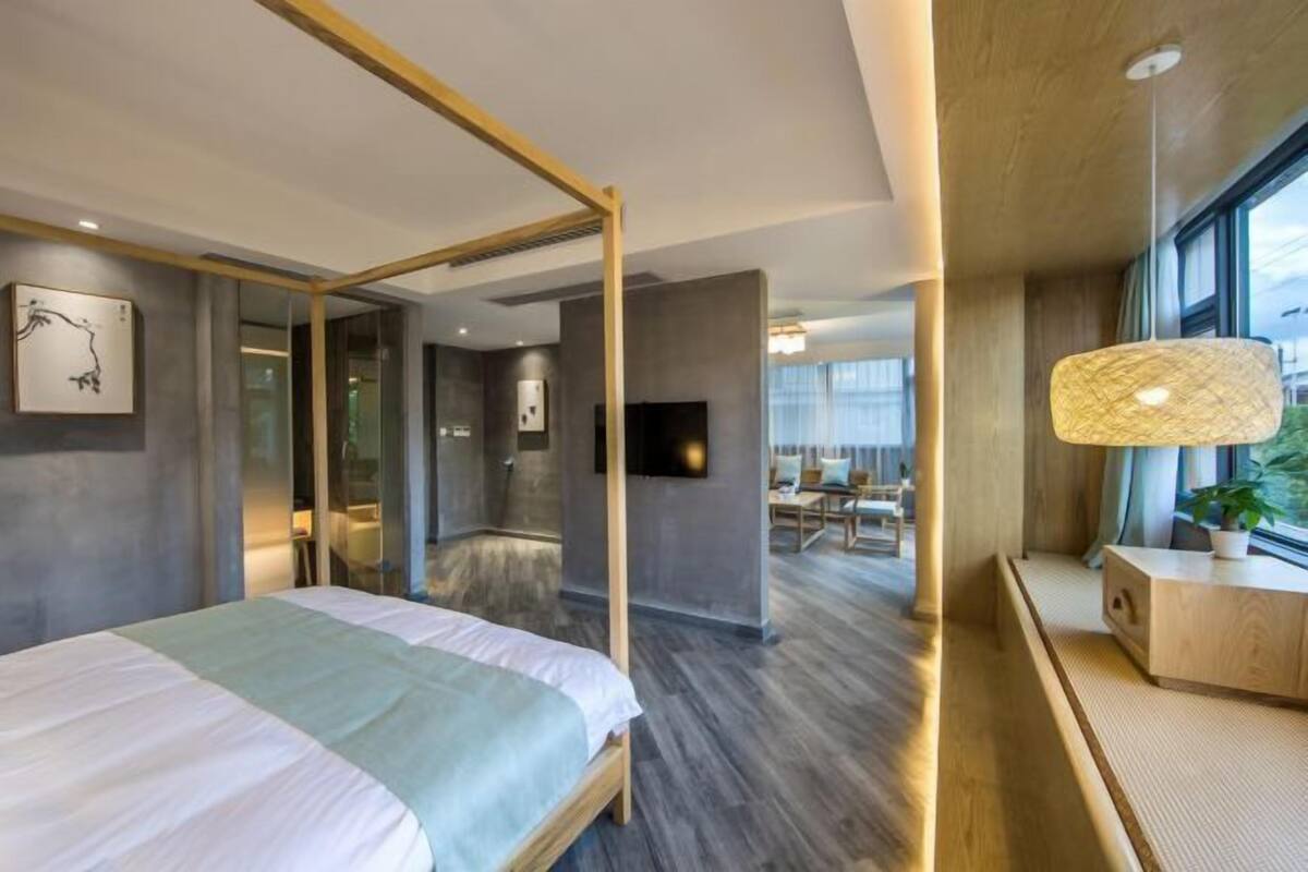 Una habitación de hotel moderna con una cama grande, un dosel con estructura de madera, un televisor de pantalla plana, una mesita de noche con lámpara y vistas a la ciudad a través de amplios ventanales.