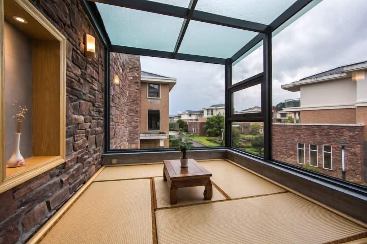 Una habitación con suelo de tatami, una mesa de madera, una planta en maceta y vistas a casas vecinas a través de amplios ventanales.