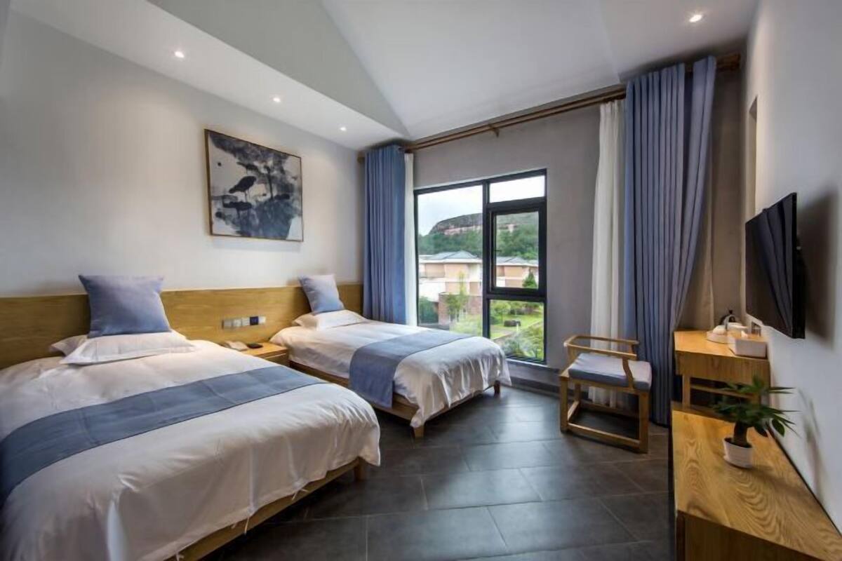 Habitación de hotel con dos camas, un ventanal grande, un televisor y una pequeña mesa con una planta.